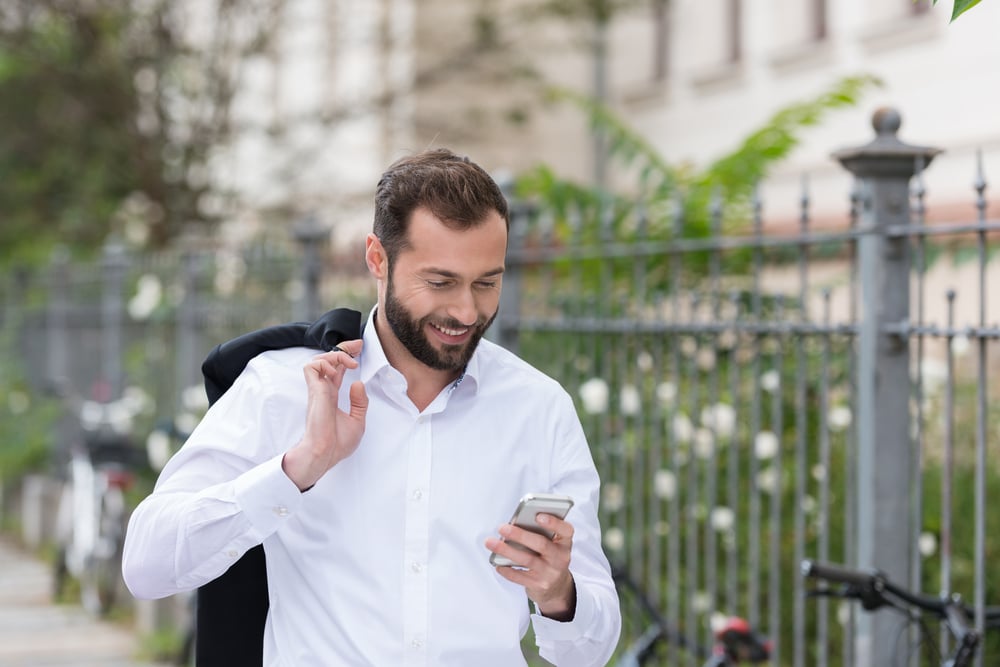 Smiling Handsome Man with Coat on the Shoulder Using Mobile Phone While Walking on the Street